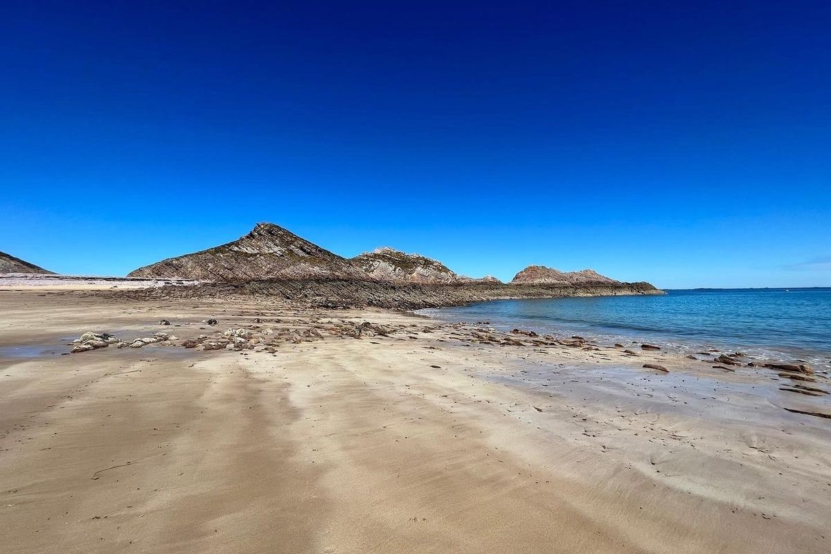 Le cap d'Erquy vu de la plage : la pointe de grès rose s'avance dans une mer turquoise, sous un ciel bleu franc