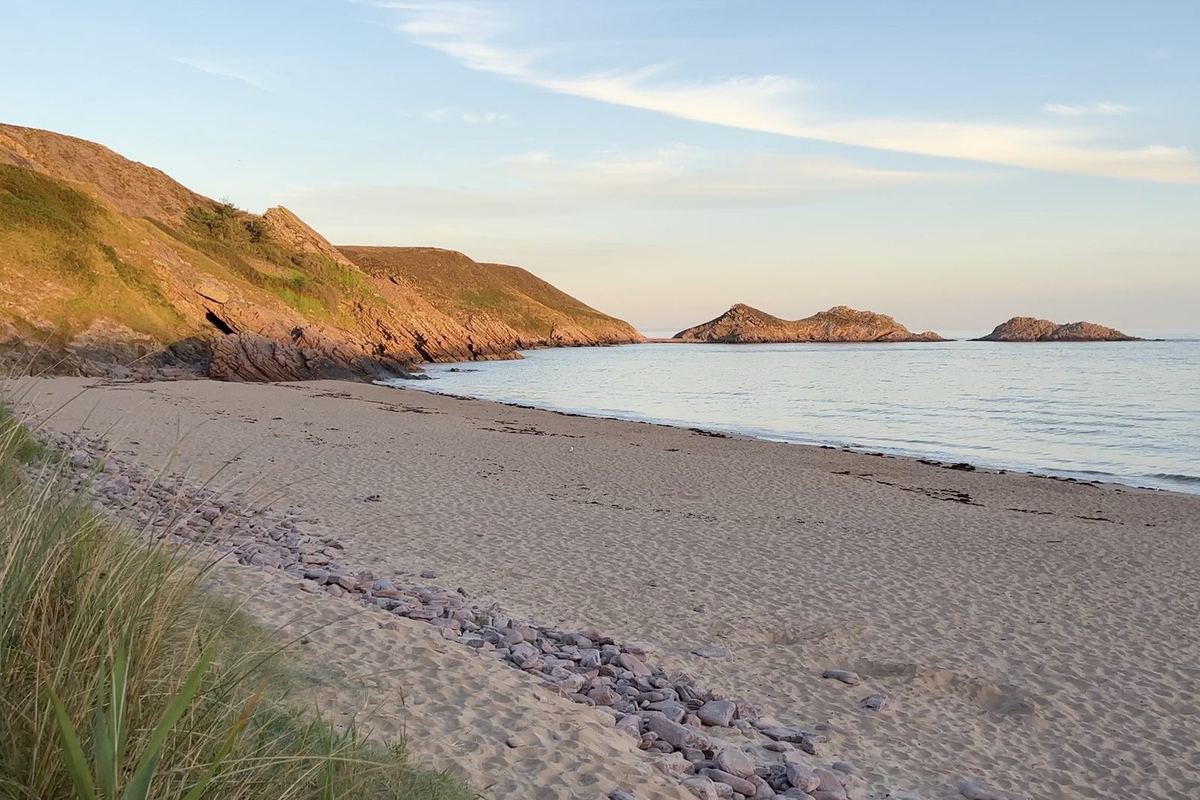 Une plage de sable au pied du cap d'Erquy en fin de journée : lumière rasante sur la falaise, mer calme et oyats au premier plan